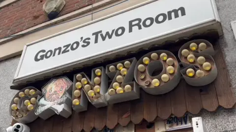 Maddy McNiven/BBC The club's entrance. Above the door, the venue's name features twice, in different styles. At the top, a white sign with black writing says Gonzo's Two Room. Underneath that the club's name appears again, this time with lightbulbs spelling out the word Gonzos.