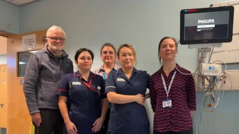 A group of five people standing next to a black screen to the right with wires coming out of the bottom, in a hospital bedroom.