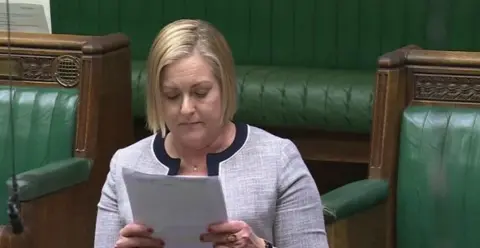 House of Commons A woman with a blonde bob-style haircut, dressed in a grey suit, reads from a piece of paper in the House of Commons.