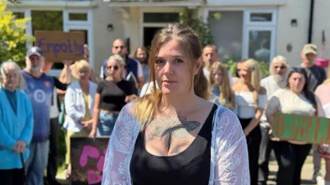 Wearing a black vest top with a thin white lace cardigan draped over her shoulders, Lucie Lambourne, with her blonde hair tied back, offers a neutral expression to the camera lens. Behind her is the house where her mum lived and a group of people including friends, neighbours and councillors, holding signs with the words 'compassion and 'empathy'. 