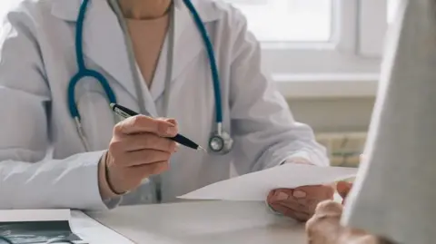 A woman GP has a consultation with a patient. She is sitting at a desk in front of a computer screen and has a stethoscope around her neck and is holding a pen and paper.