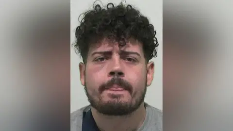 Mugshot of Kier Adam-Nuhoglo. He has thick black curly hair, a black beard and a reddened face. He is sticking his tongue out slightly between almost closed lips.