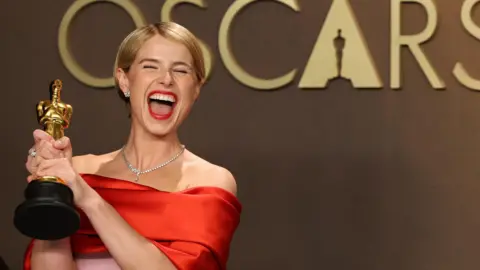 Jessie Buckley with her eyes closed smiling widely at the camera. She is holding an Oscar award and is wearing a red and pink satin dress.