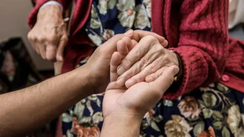 An elderly person's hand being held by another person's hands. The elderly person is wearing a flowery dress and red cardigan, but their head is not in the image