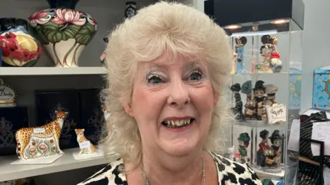 Kathy Loudon smiles into the camera. She has silver eye shadow, pink lipstick and permed blonde hair. She is standing in front of two display shelves covered in ornate pottery.