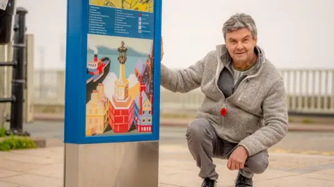 Alan Roper crouching next to his artwork on a signage monolith in Maryport.