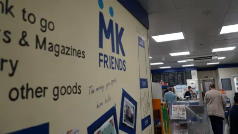 In the foreground is a noticeboard or advertising board, with "MK FRIENDS" branding in blue font printed on it. In the background, there are some people queuing at a counter in the MK Friends shop. Behind the counter on the wall is a menu or price list chalked on a black board.