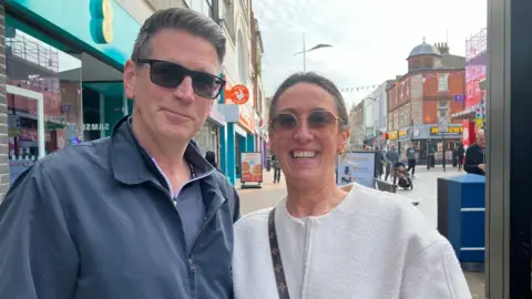 Henry Godfrey-Evans/BBC Peter and Tammy Allen, a man and a woman, are standing in a high street. The man is wearing a dark blue jacket and sunglasses. The woman is wearing a white top and sunglasses.