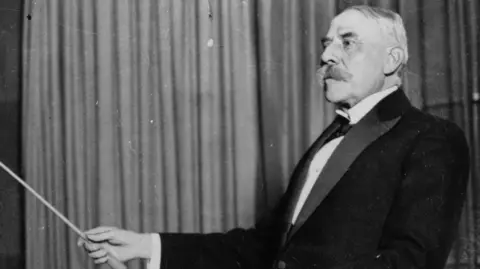 Getty Images Composer Sir Edward Elgar conducting with a baton. He is on the right in this black-and-white image.