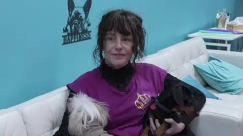 A woman in a purple top and black sleeves is sitting on a white leather sofa holding two dogs