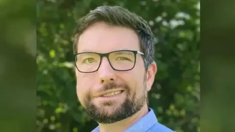 Aaron Jones looking at the camera. He is wearing a blue shirt and dark-rimmed glasses and dark hair. He has a short trimmed beard and moustache