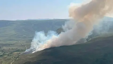 A green hill landscape with plumes of white smoke billowing into the air