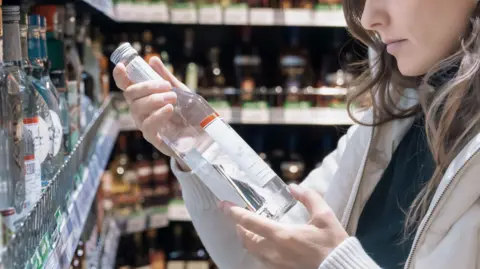 In a supermarket, a brown haired woman carefully inspects the label on a vodka bottle in the alcoholic beverage aisle, deciding on her purchase. 