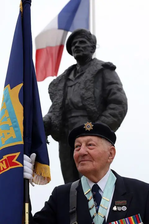 Matt Cardy/Getty Images Normandy veteran Mervyn Kersh stands below a statue to him at a memorial service close to the British Sword beach at Colleville Montgomery on June 5, 2013 near Caen, France. He is wearing a suit and tie with medals on his jacket. He is holding a flag pole which has a navy blue flag with yellow and red sections on it. The flag of France is seen in the background.