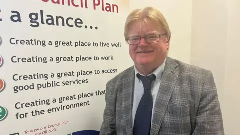 Man with blonde hair and glasses in grey jacket and navy tie stood infront of a advertising board for a local council