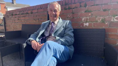 BBC/Lucy Ashton A man with grey hair is smiling. He is wearing a light grey blazer, a lavender coloured tie, a shirt with large checks and pale blue trousers