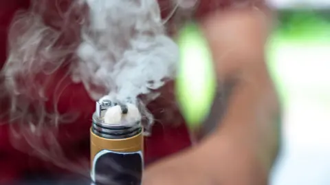 A close-up picture of a vape with smoke coming out of it. It is being held by a man who is very blurred in the background. 