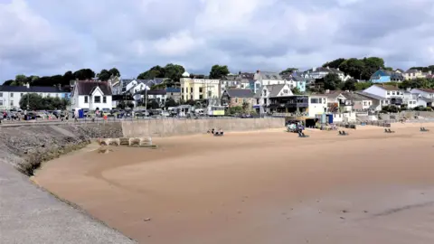 Oscar Hill/ Getty Images This image shows Saundersfoot beach, the sand is beige and the sky is blue. There's a stone wall separating the sandy beach from a collection of brightly coloured buildings. 