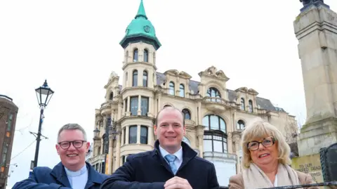 Standing in front of the Austins building in Londonderry, are Minister Gordon Lyons, Rev Robert Miller and Helen Quigley from the Inner City Trust.