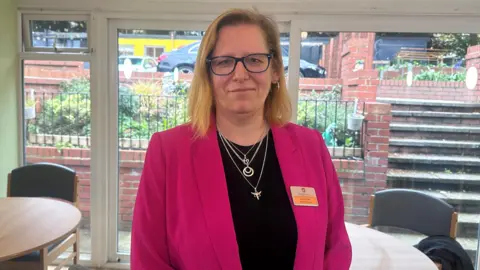 Alena Petrie is standing in front of a large window overlooking the garden and car park. There are a number of tables and chairs dotted around the room. 
