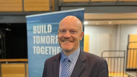 Gavin St Pier pictured wearing a dark coloured suit jacket, a grey-ish shirt and a blue and white polka-dotted tie. Mr St Pier has stubble and a bald head.