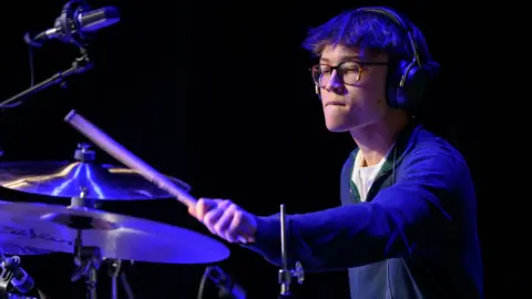 Ellis Gates wearing headphones and glasses and playing the drums. He is wearing a blue jumper
