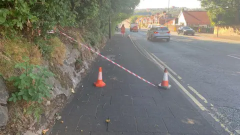 A pavement and road in West Road in Newcastle. There are two cones with some orange tape stretching across it. 