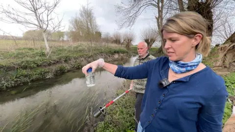 Jo Robb, green councillor for South Oxfordshire District Council, is holding a small bottle of water near the brook. A man behind her is holding a pole. It is cloudy. The reeds can be seen in the water.