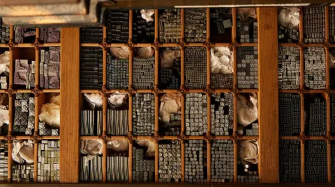 Jess Hand Photography A wooden tray pictured from above, with lots of individual rectangular slots for metal stamps of symbols and letters.