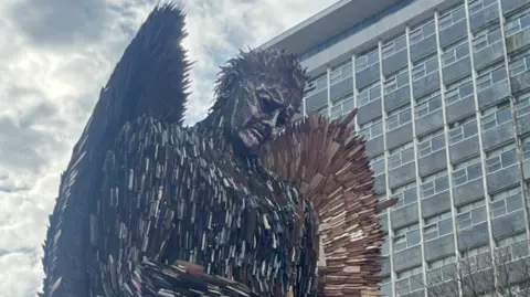 A side view of the Knife Angel. It has a sad facial expression and is looking down. The knife handles are visible of its body, while large, rusty blades poke out of its wings. A large grey building is behind the structure.
