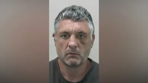 Northumbria Police Musghot of William Flamson. He has faint grey stubble and short thick dark grey hair. He has a bruise under his left eye and a faint scar on his right cheek.