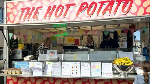 Caring Cooks The image shows a food truck called THE HOT POTATO, decorated in red and yellow with potato-themed graphics. Inside the truck are three people and one customer in front. The counter displays a bowl of bananas, printed materials, and other food items. 