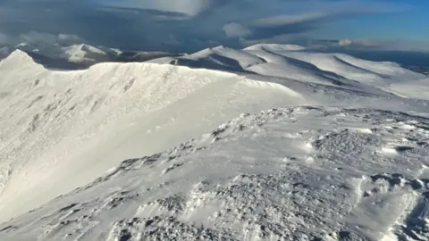 Summit of Blencathra in the Lake District. A snow-topped summit with blue sky and dark clouds in the distance. 