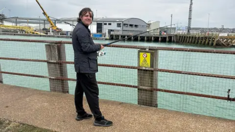 A man in a black coat and black trousers is fishing by a harbour. 