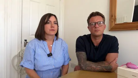 A brunette woman in a striped white and blue shirt. She is wearing a gold necklace. She is sitting next to a man, with dark hair and wearing a black t-shirt and tattoos on both his arms. 