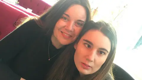 Two women both with long dark hair and dressed in black pose together. The older woman on the left smiles and wears a gold necklace. The younger woman on the right looks serious and distracted.