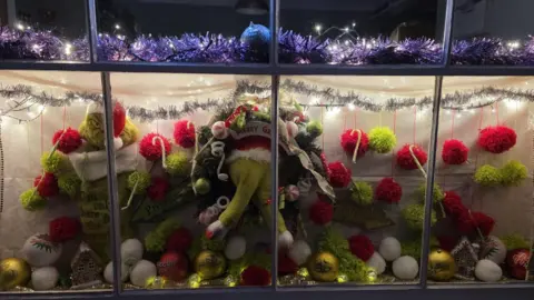 The window is full of a Grinch cuddly toys and red and green pompoms. There is a banner which reads "Happy Grinchmas". There is tinsel and fairy lights above the scene.