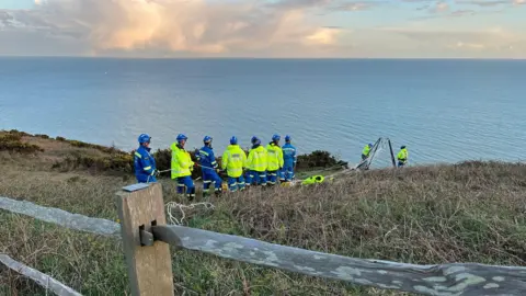 Coastguard teams in high‑visibility jackets and helmets stand on a grassy clifftop. Some are near the edge with rope equipment, while others are further back. In the background is a wide expanse of sea under a pale blue sky with scattered clouds. A wooden fence runs across the foreground.