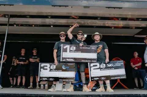 Dale Wherton Three man are standing on a stage, all wearing back tshirts and shorts and caps, they are all holding a sign each, which say 3rd, 1st and 2nd place. 
