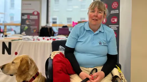 Jenny sits in a chair looking at the camera. She has a light blue polo t-shirt on with a Guide Dogs logo on it with a black underlay and cream coloured trousers. She has a lead in her hands and her Labrador Retriever sitting to her side which is looking to the left of the photo.
