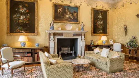 A drawing room with pale yellow patterned wallpaper, a white marble fireplace at the centre, and three large framed paintings above it. In the foreground are matching upholstered sofas and a round table on a floral carpet, with bookshelves, ceramic lamps, and decorative plates arranged along wooden cabinets.