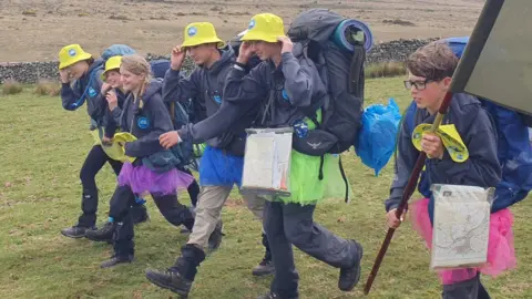 Six children from the scouts are walking next to each other wearing hiking gear and carrying backpacks. They are in a field at the start of the Ten Tors challenge. 