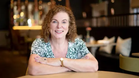 Karen Hester with curly ginger hair wearing a green and white top with a gold watch. Her arms are crossed and she is leaning on a table while looking into the camera.