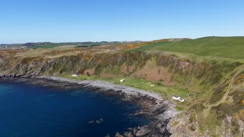 Galbraith A view across water to cliffs with a small number of cottages at the bottom. In the distance is the nearest village