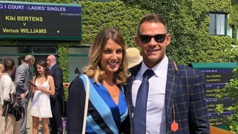 Andrea Byrne / Y Lolfa Andrea, dressed in a blue dress, stands next to Lee, wearing a blue suit, smiling at Wimbledon 