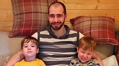 Barry has short dark receeding hair. He is wearing a black and with stripey t-shirt and is sitting on a sofa with his two sons who both have short brown hair. One is wearing a yellow t-shirt, the other a grey top. There are red cushions in the background.