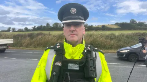 Ch Supt Norman Haslett is standing on the side of a road. He is wearing a PSNI cap and a yellow high viz jacket with a body vest. There's a walkie-talkie attacked to his vest. He has medals on his lapels. Fields are in the background. Some cars are passing. A tripod with a camera attached is to his side. 