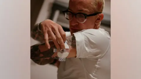 @bfarbr Jonty Brown is wearing a white shirt and is attaching a blood glucose monitor, a white round device, to the back of his arm