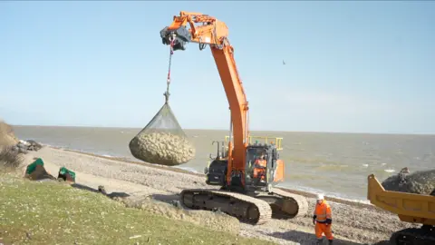 A large piece of machinery loads sea rock bags on to a coastline. Workers in orange hi-vis walk around the beach nearby. 