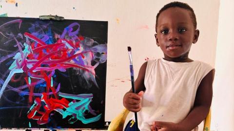 A toddler holding a paintbrush in front of a painting 